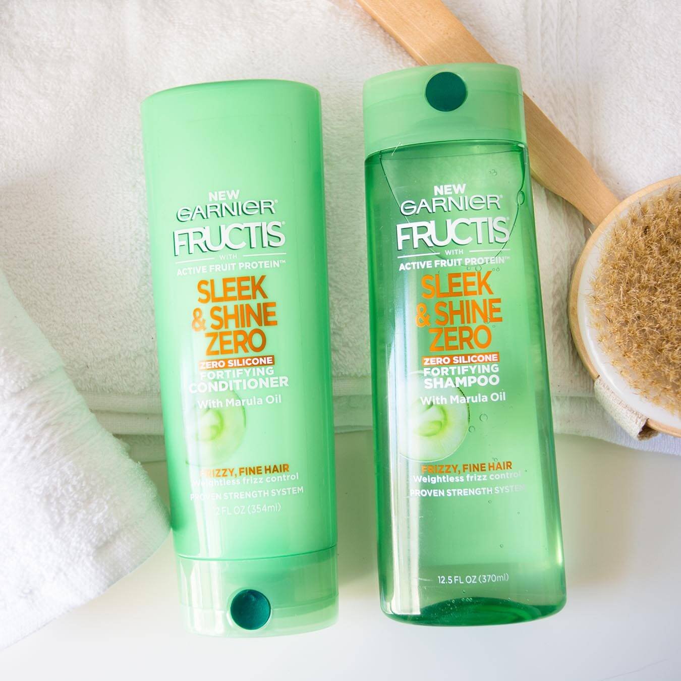 Garnier Fructis Sleek & Shine Zero Shampoo and Fructis Sleek & Shine Zero Conditioner on a white bath towel on a white background next to a white hand towel and a wooden bath scrub brush.